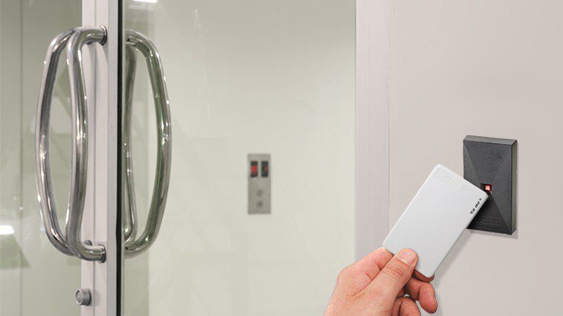 Hand holding a white access card near a wall-mounted card reader by a glass door