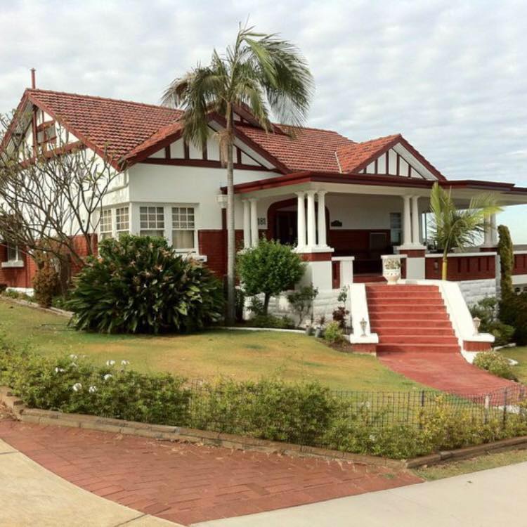 Large white house with red roof, front porch, palm trees, and a red brick driveway