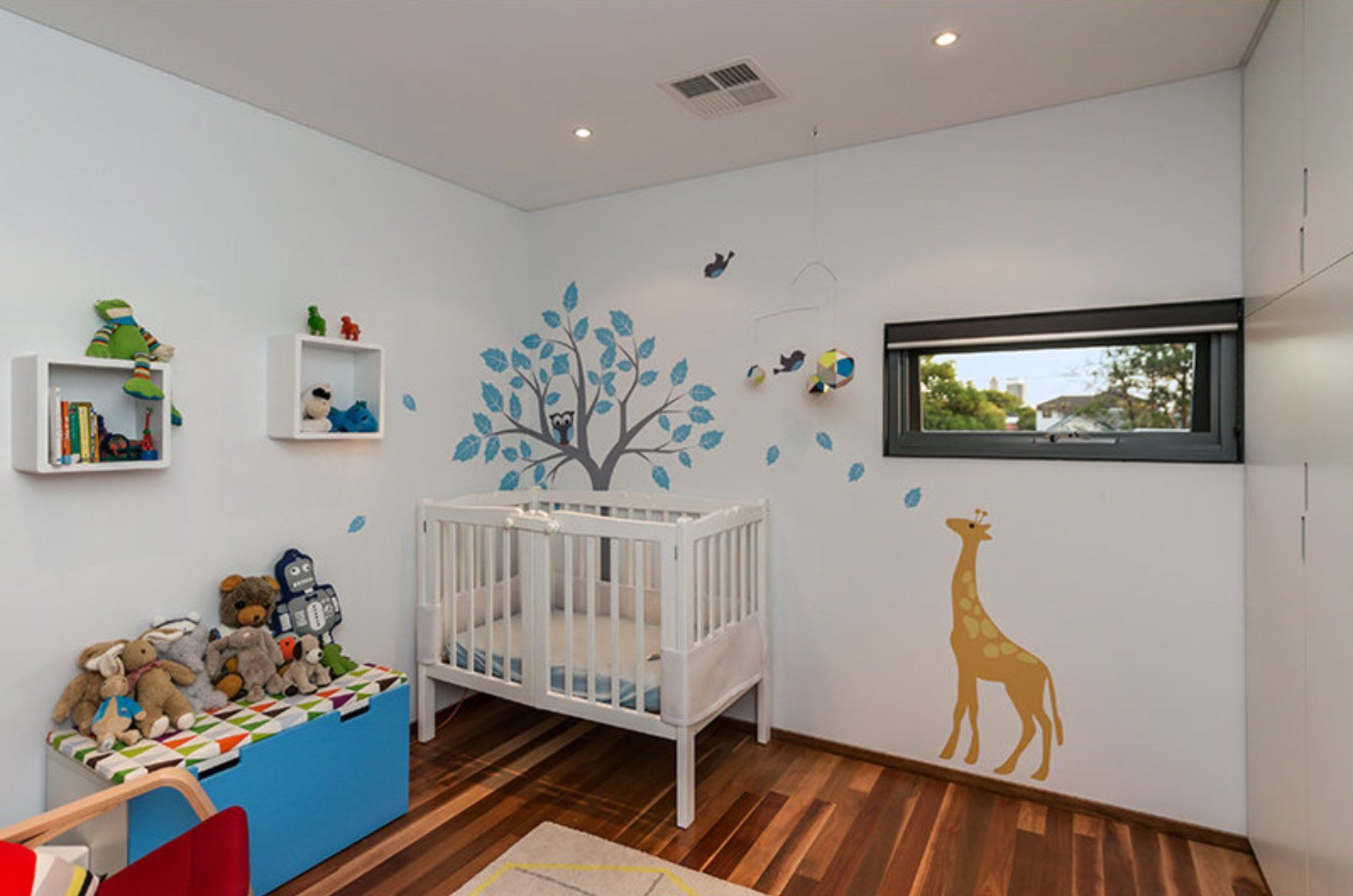 Bright nursery with white crib, blue tree wall decal, yellow giraffe decal, and toys by a window