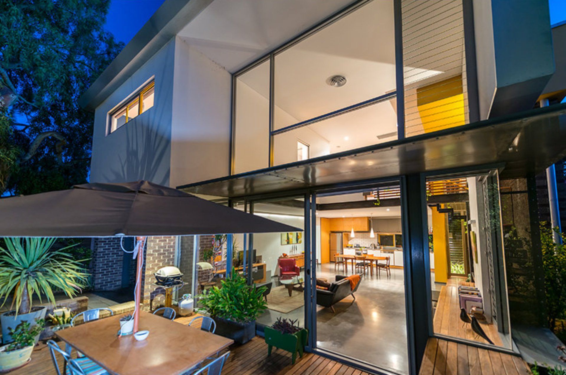 Modern two-story glass house at dusk with a lit patio and indoor dining area visible through large windows
