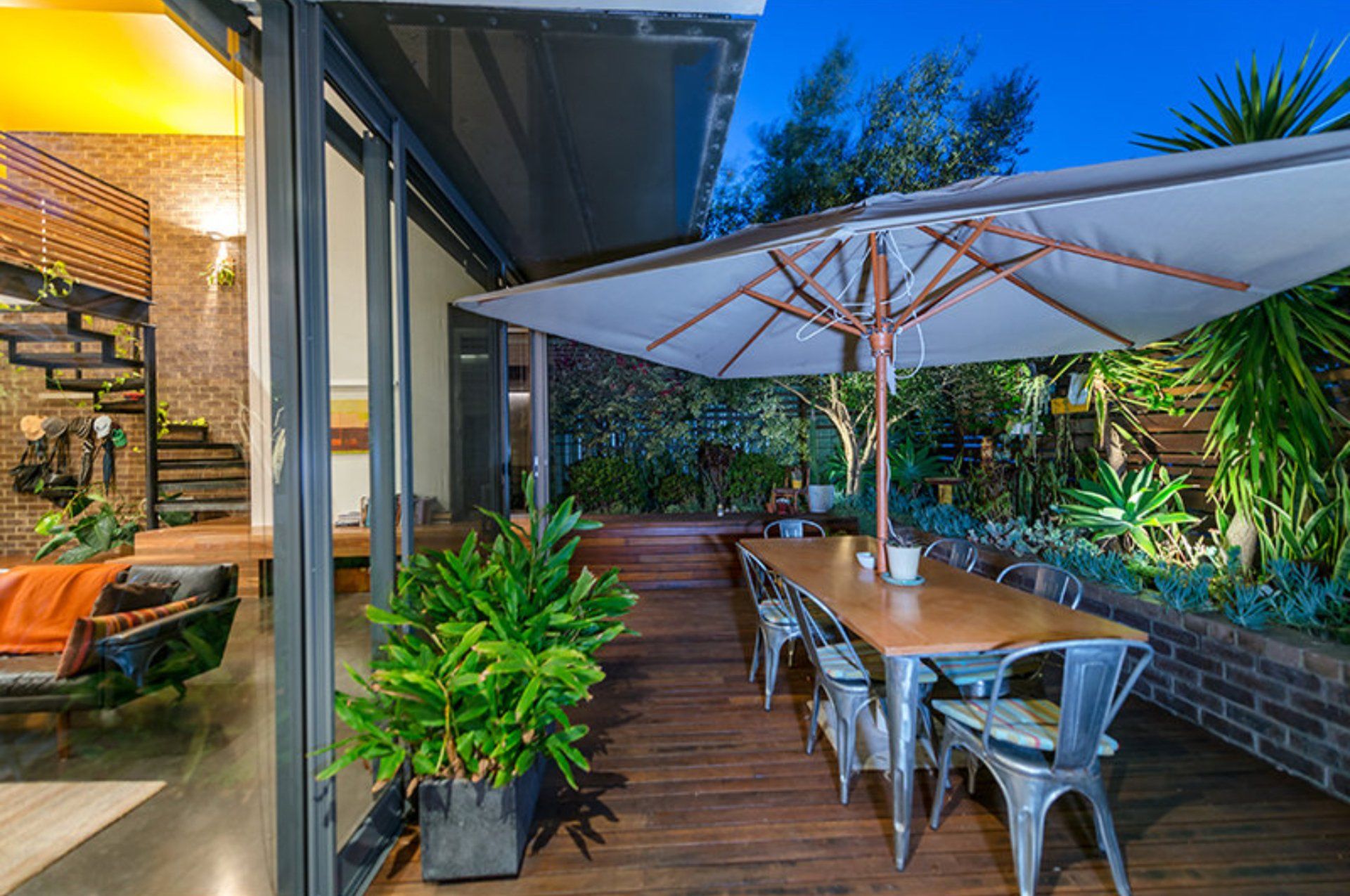 Outdoor patio with wooden deck, metal table and chairs, and a large white umbrella beside lush greenery