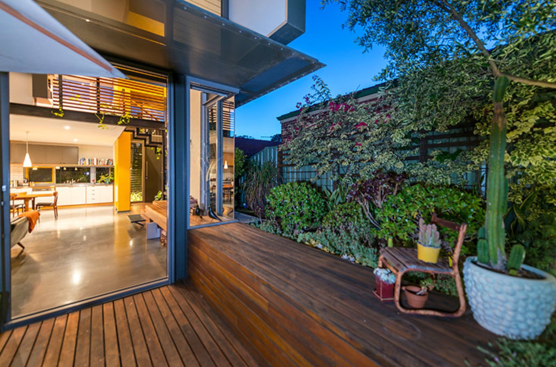 Modern home patio with wooden deck, open glass doors, warm indoor lights, and potted plants beside a green wall