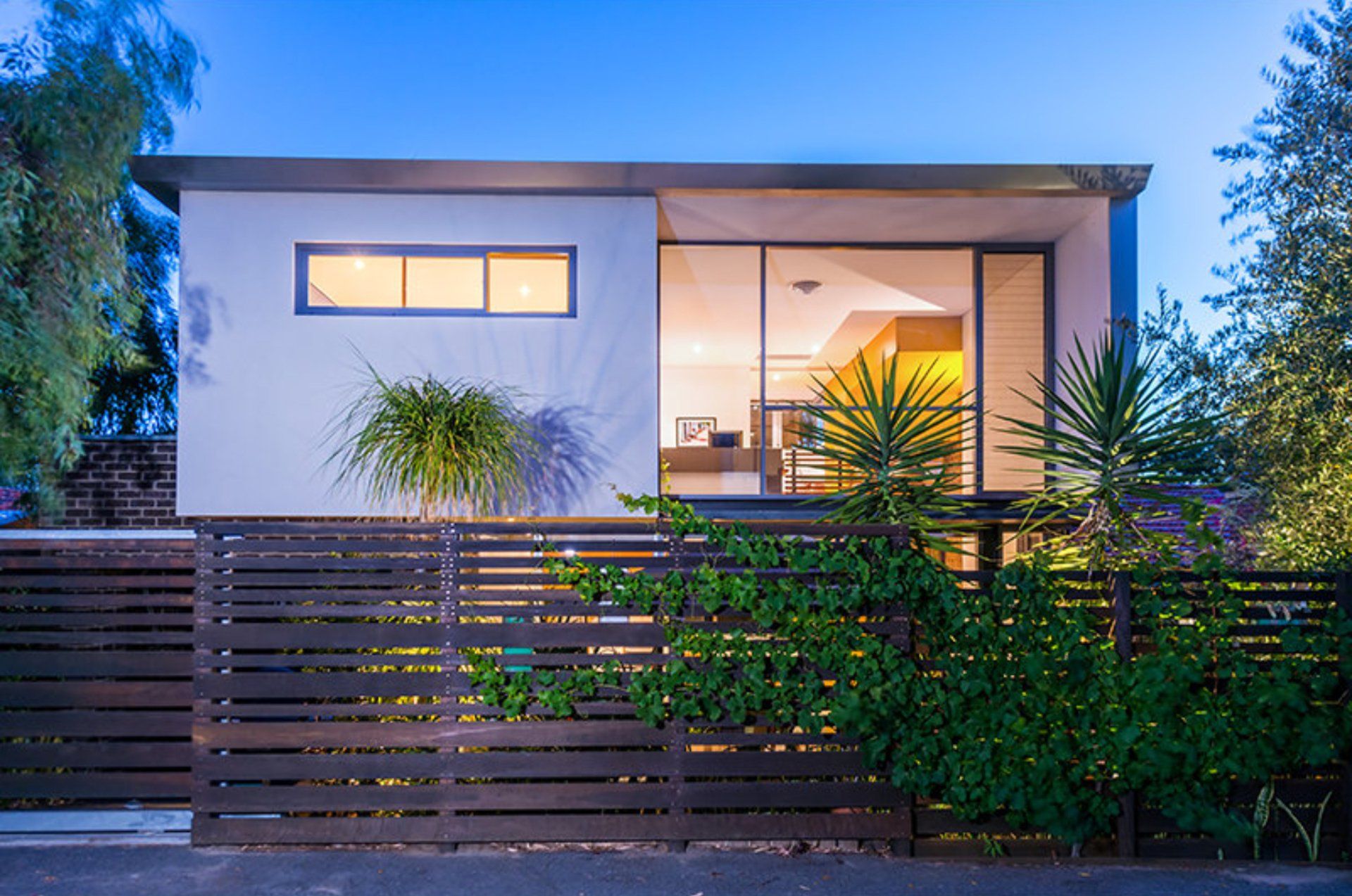Modern white house with lit windows, wooden slat fence, and lush greenery at dusk