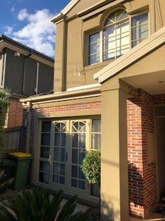 Two-story tan house with brick porch, large windows, and a small front garden