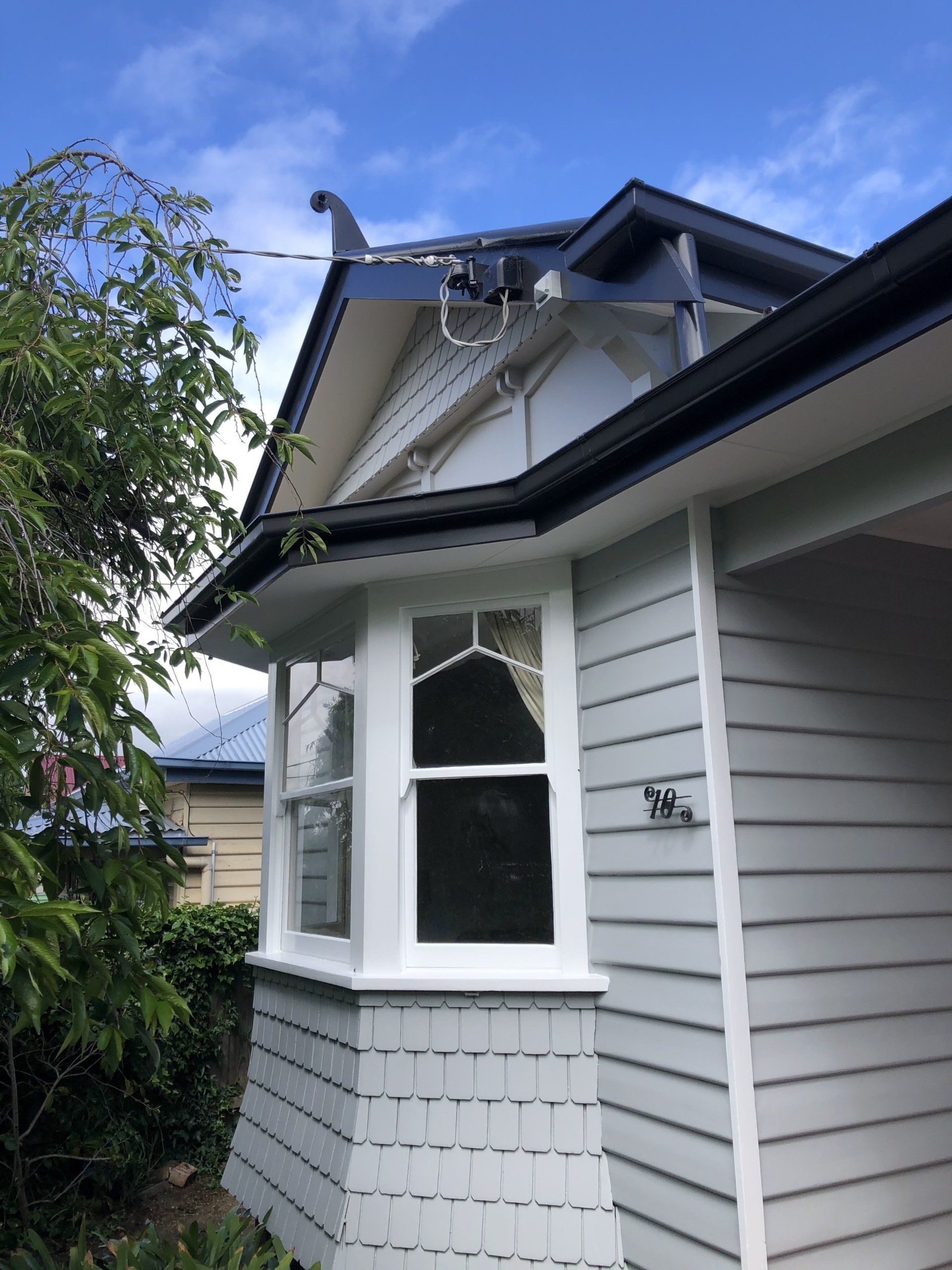 White house exterior with damaged roof and a broken gutter under repair, beside a leafy tree.