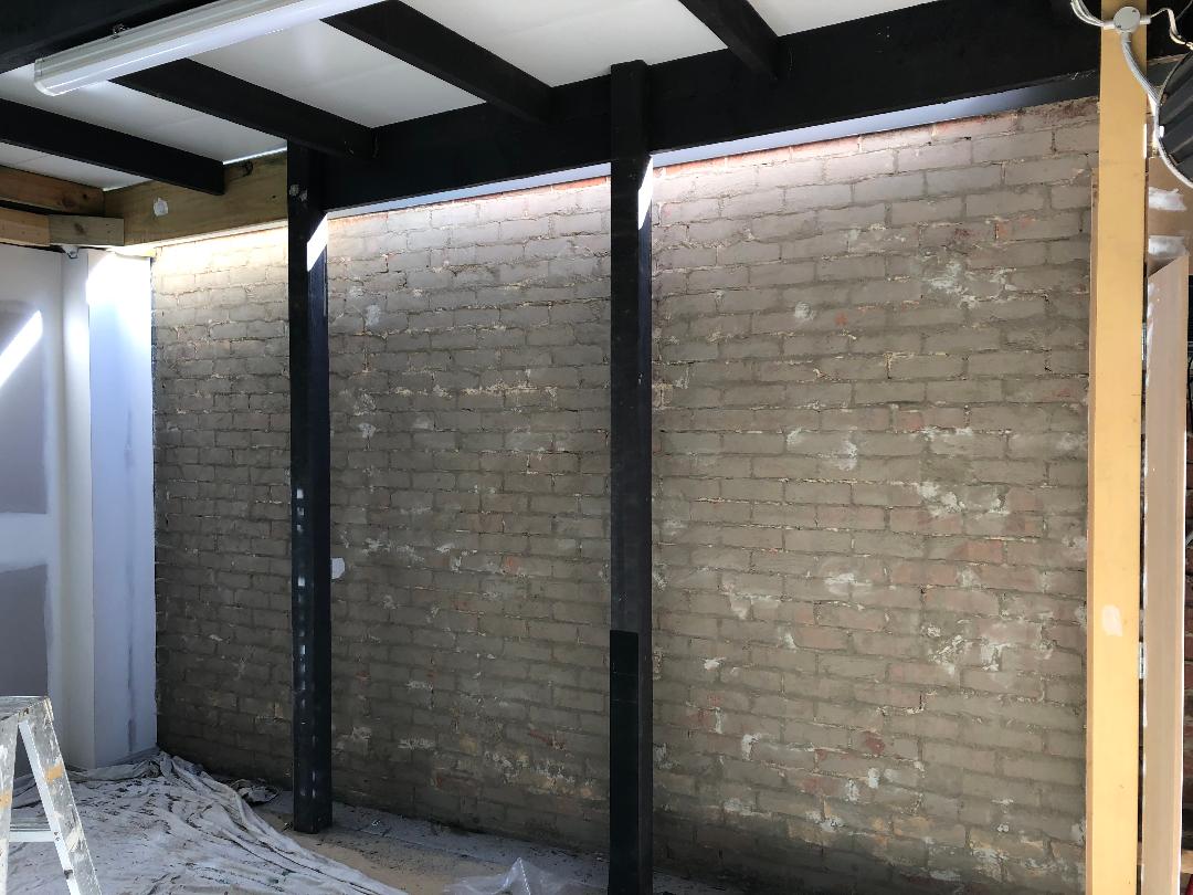 Exposed brick wall with black steel beams in an unfinished room, with debris on the floor and a ladder at left