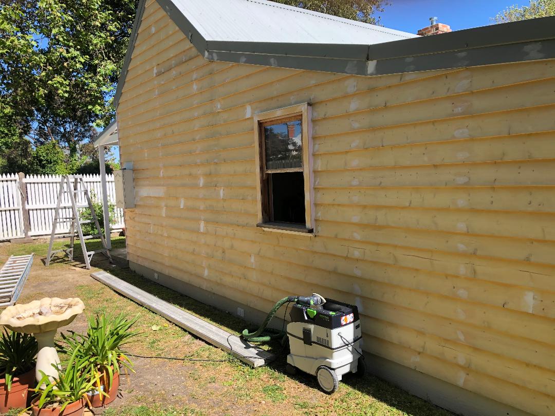 Side of beige house with a small window, yard tools by the wall, and a fenced garden in sunlight