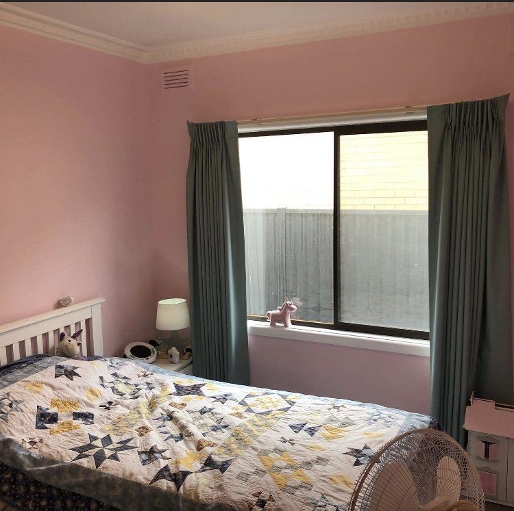 Pink bedroom with a bed, dark curtains, and a large window letting in daylight
