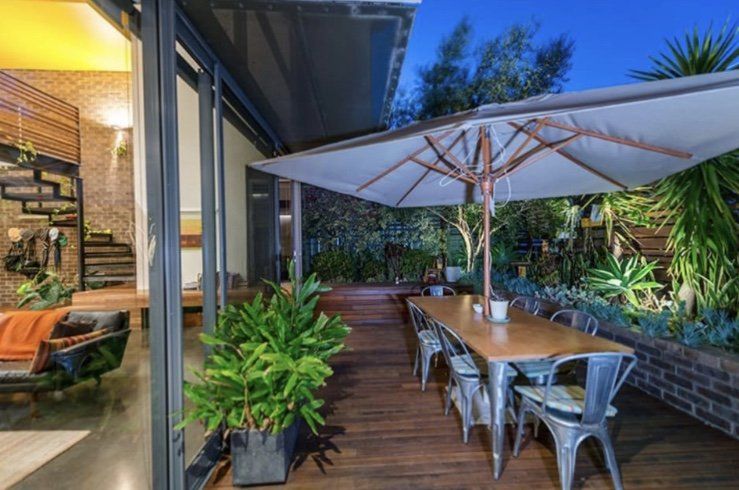 Outdoor wooden patio with dining table, metal chairs, large umbrella, and lush plants beside a house at dusk