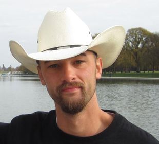 A man wearing a white cowboy hat stands in front of a body of water