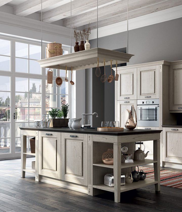 Cucina dai colori chiari con isola e portapentole sospeso. Ampie finestre, ripiani scuri e pavimenti in legno.