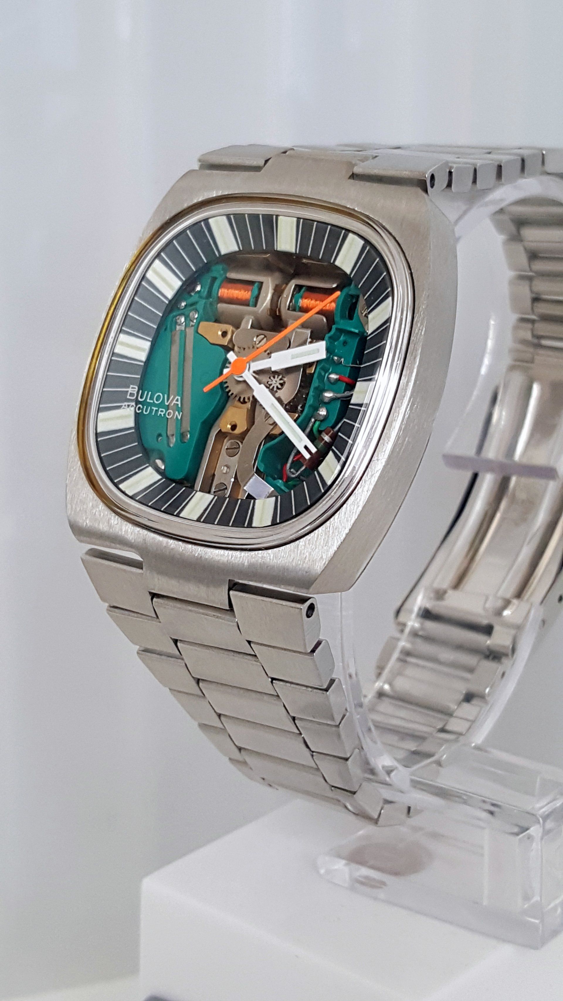 Silver wristwatch with a teal skeleton dial on a clear display stand