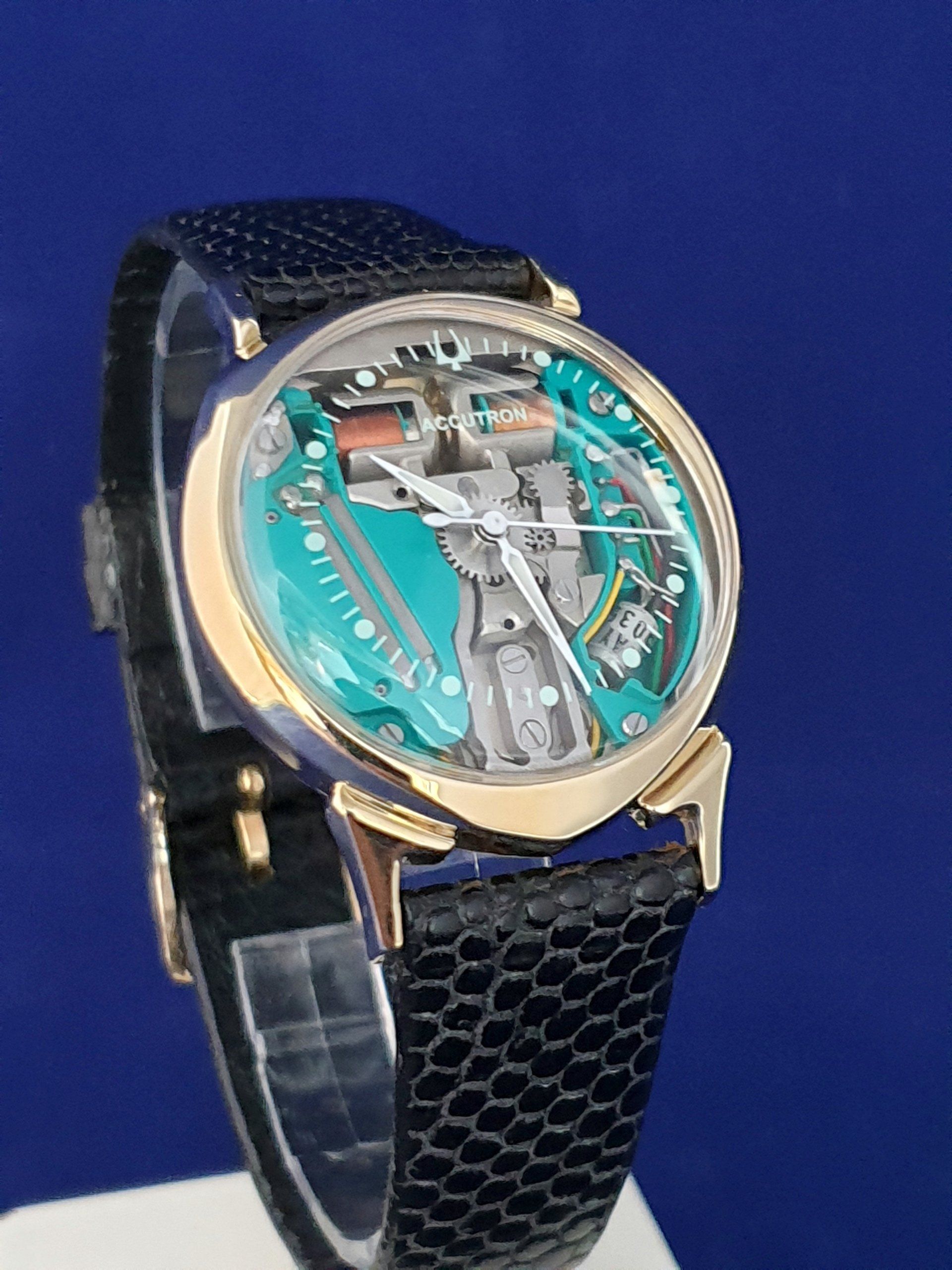 Luxury wristwatch with gold case, turquoise skeleton dial, and black textured band on a blue background