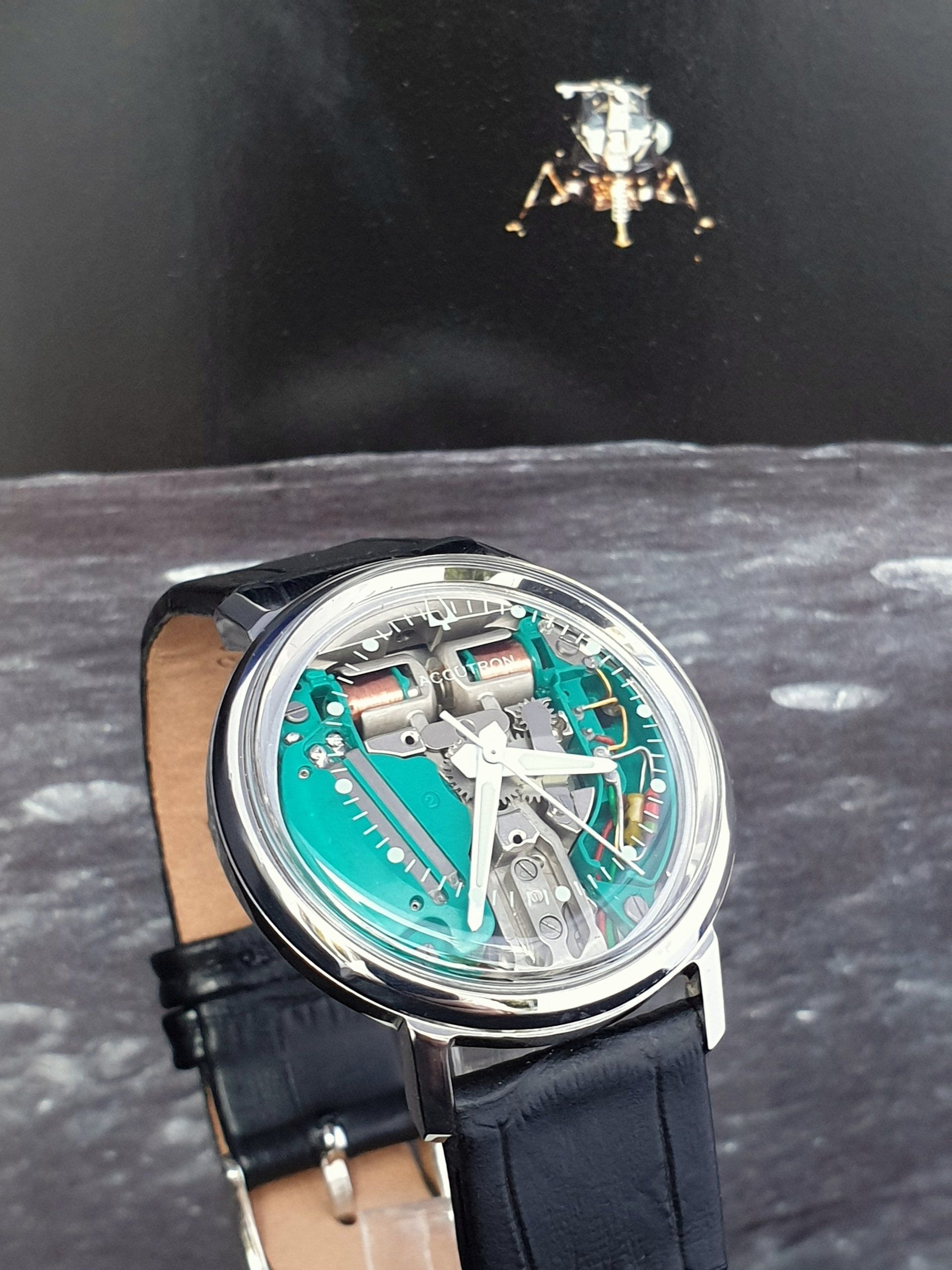 Luxury wristwatch with black strap on a wrist, against a dark space-themed background with a small spacecraft.