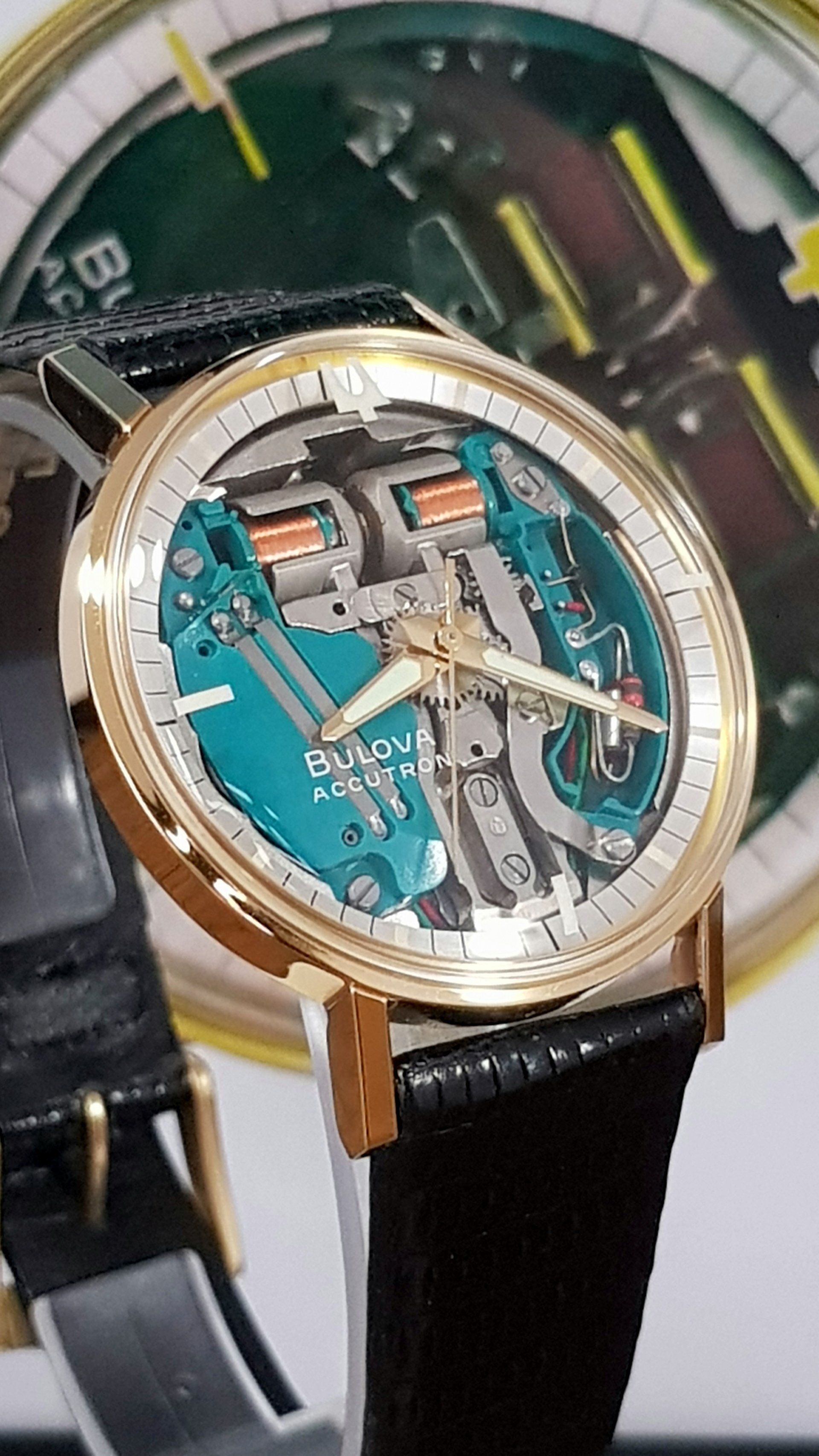 Close-up of a teal skeleton wristwatch with a gold bezel and black strap, shown against a blurred watch background