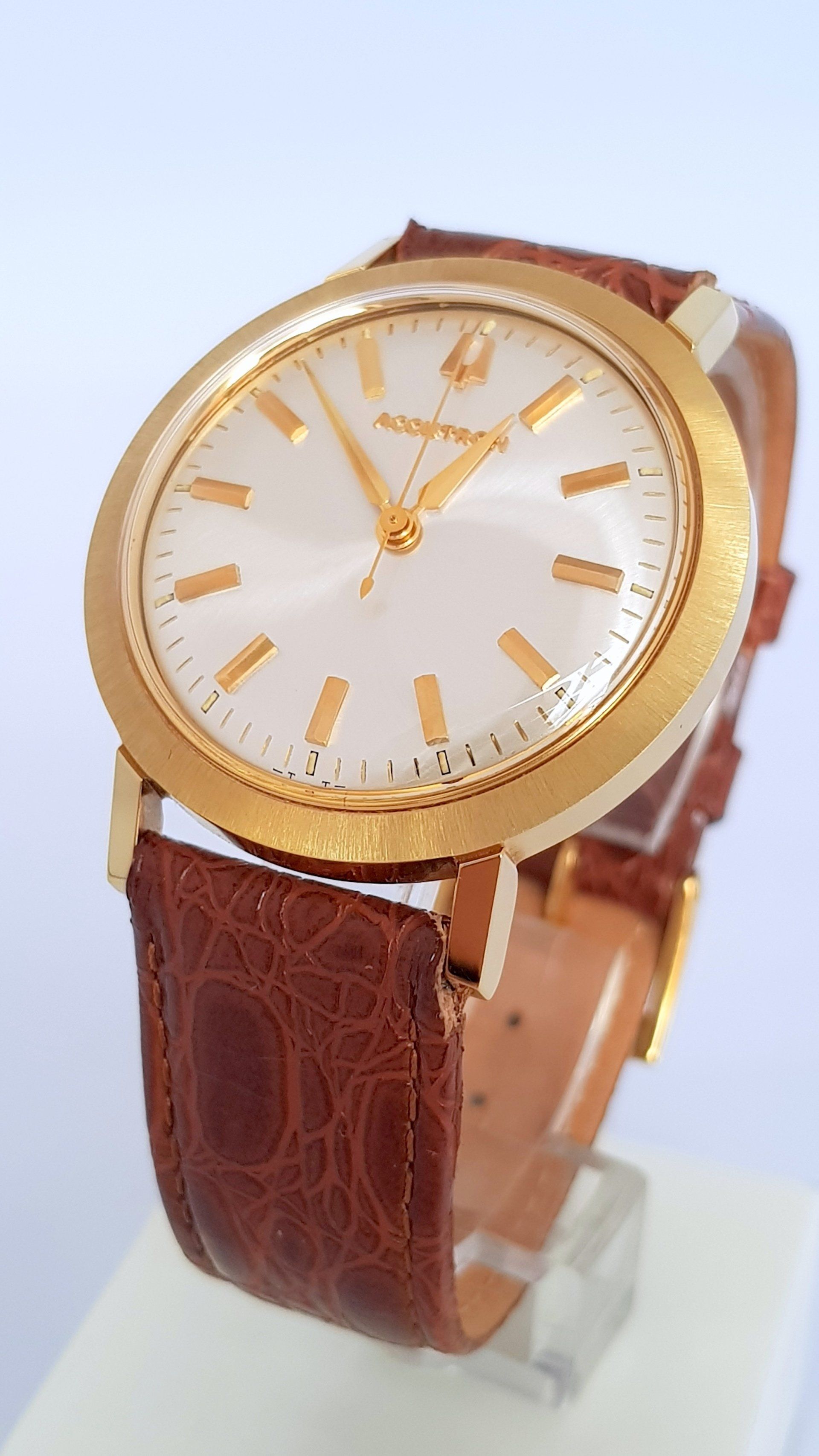 Gold wristwatch with white dial and brown leather strap on a clear stand