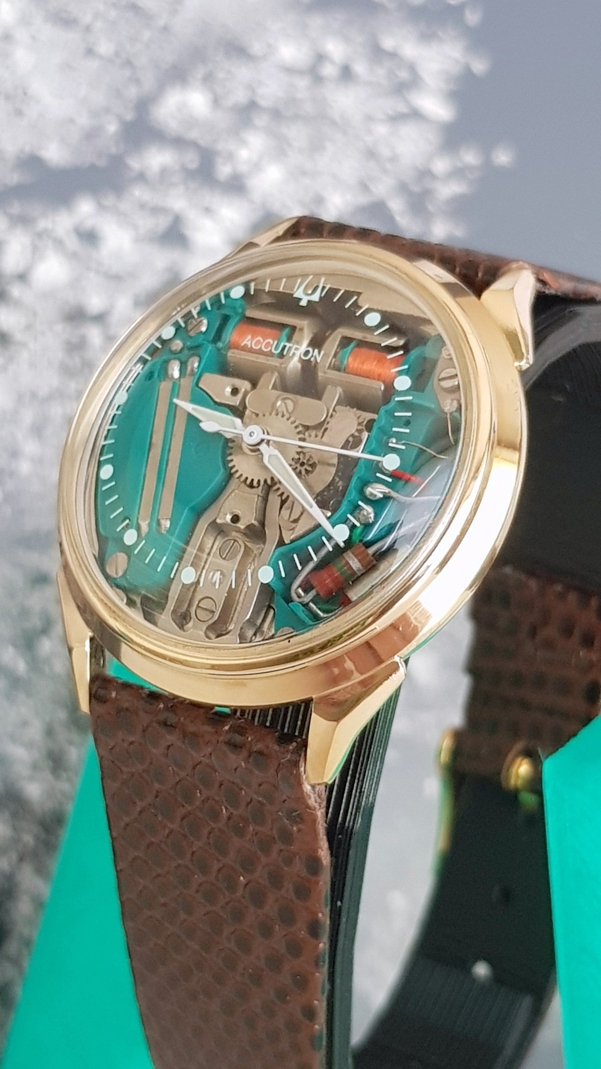 Gold watch with turquoise skeleton dial and brown leather strap against a cloudy sky