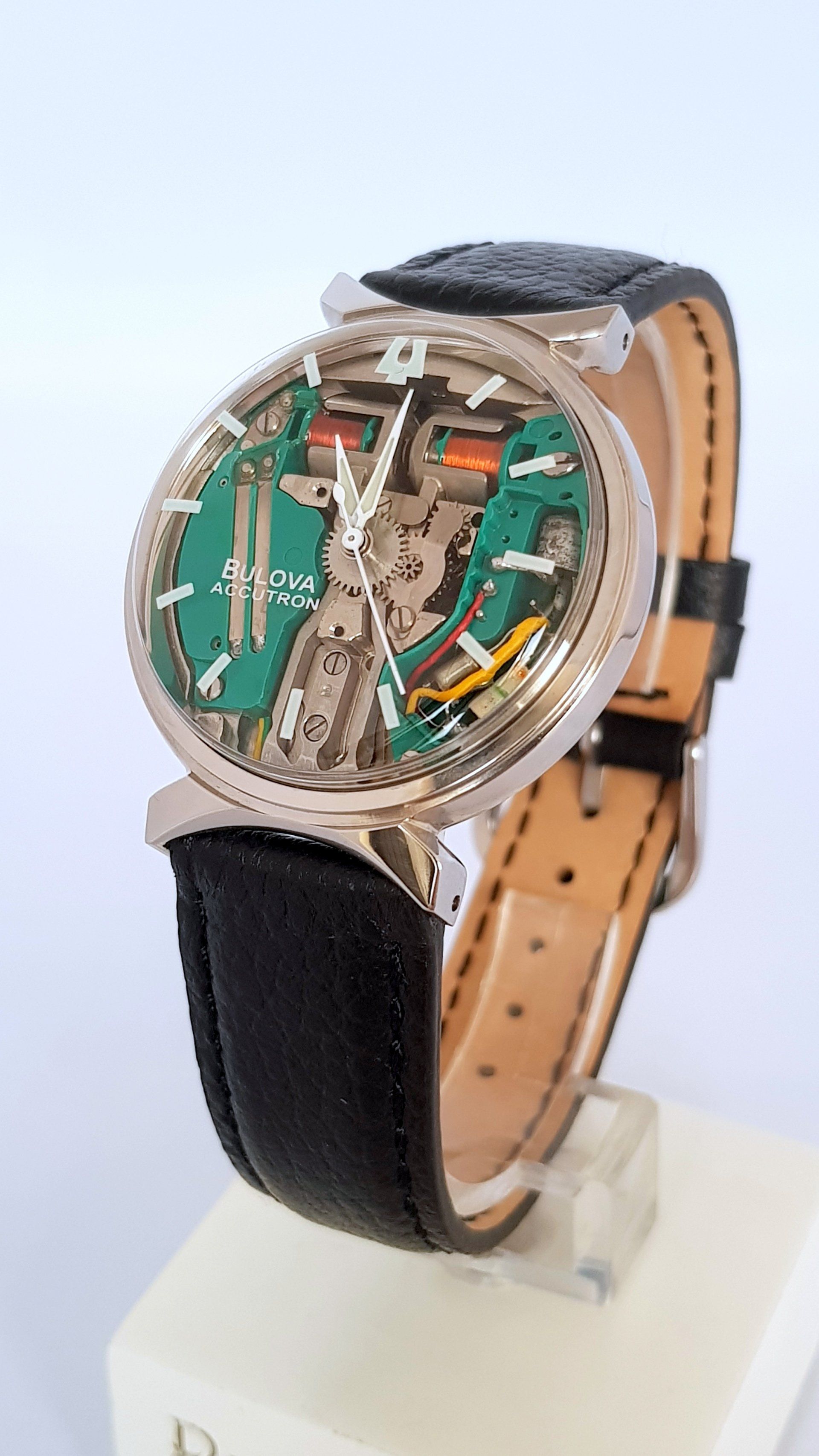 Skeleton wristwatch with green dial, tan leather strap, and black band displayed on a stand