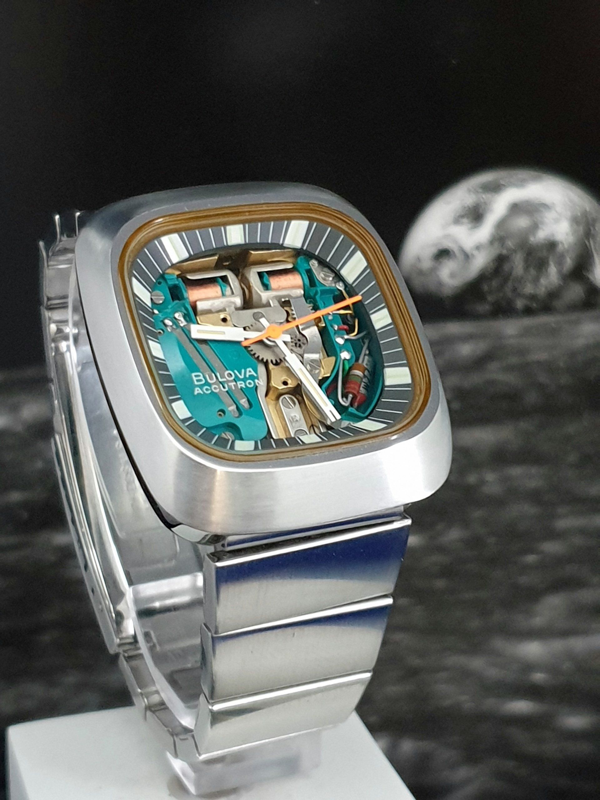 Silver luxury wristwatch with turquoise dial and gold accents on a clear stand, against a dark background