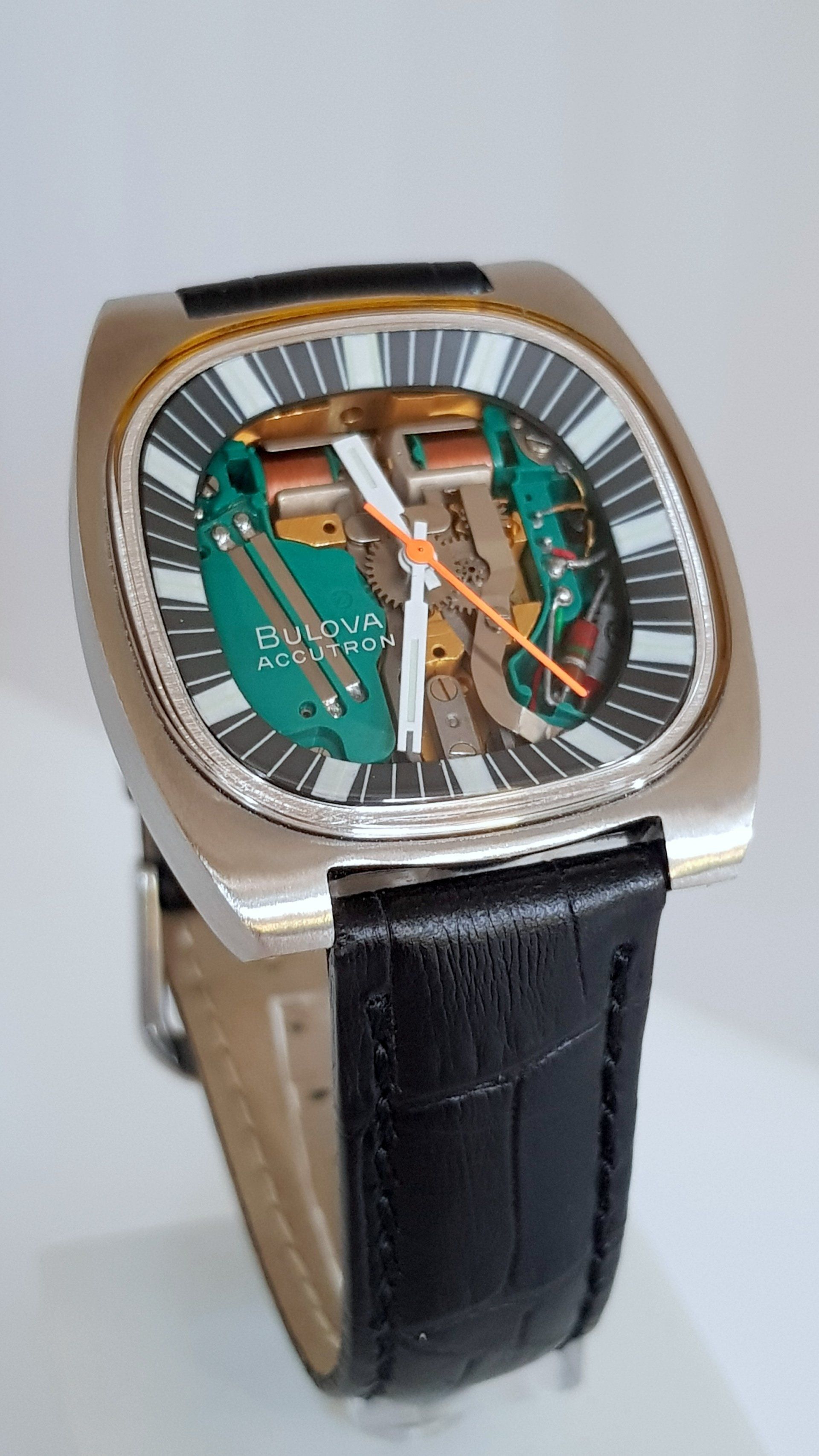 Luxury rectangular wristwatch with silver case, skeleton green dial, orange hands, and black leather strap