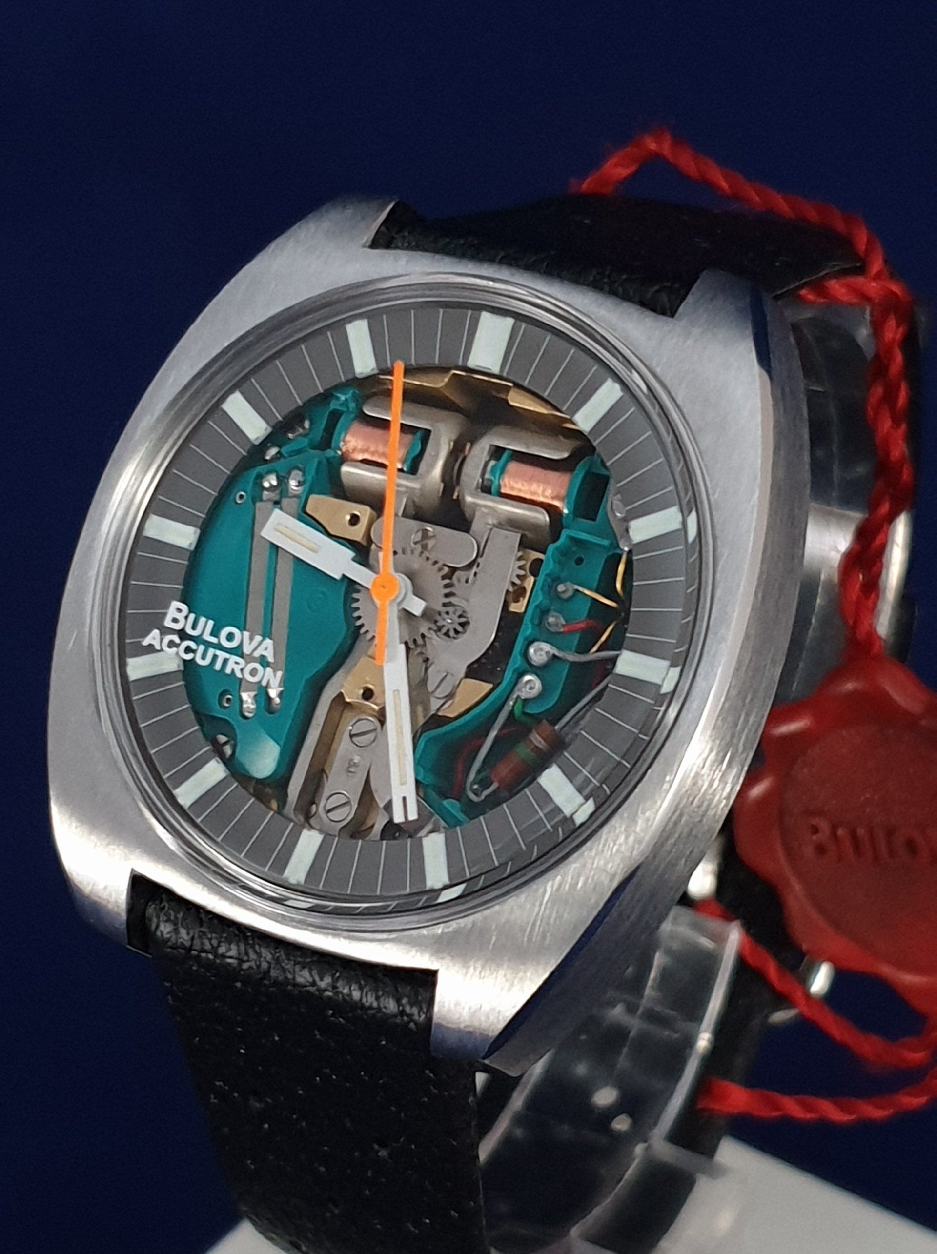 Close-up of a silver and teal mechanical wristwatch with a black strap and red cord on a dark blue background