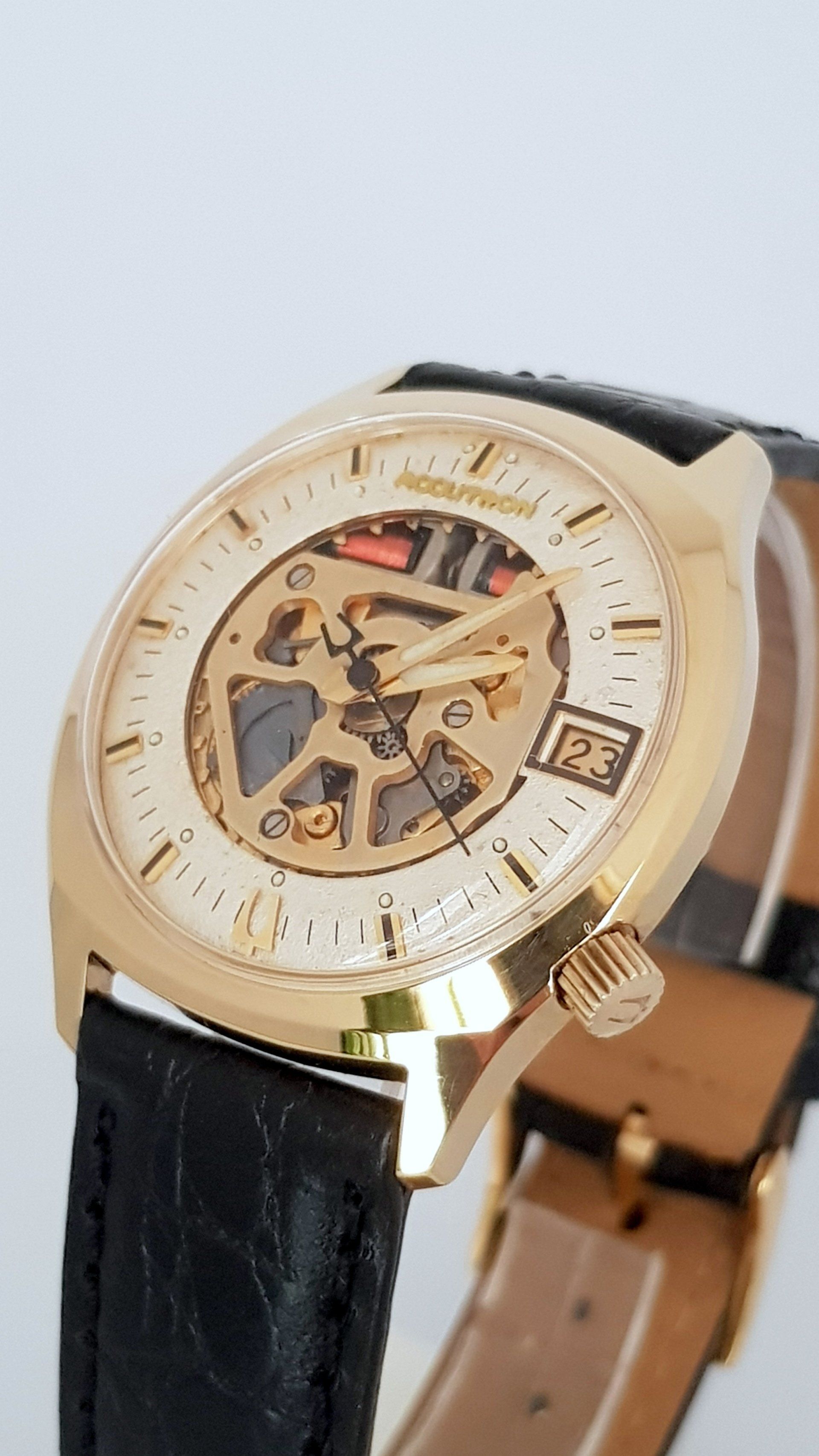 Gold skeleton wristwatch with black leather strap on a light background