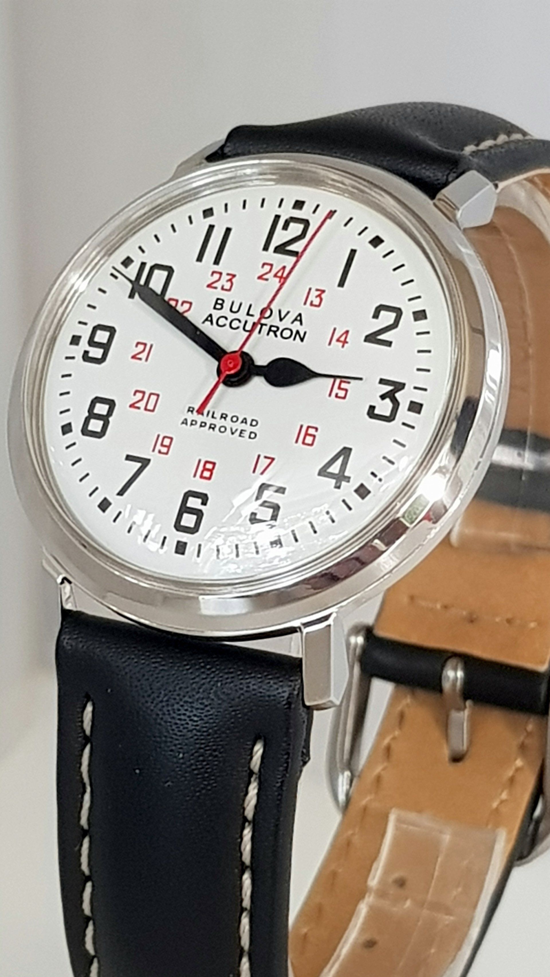 Silver wristwatch with white dial, black numerals, and black leather strap on display stand