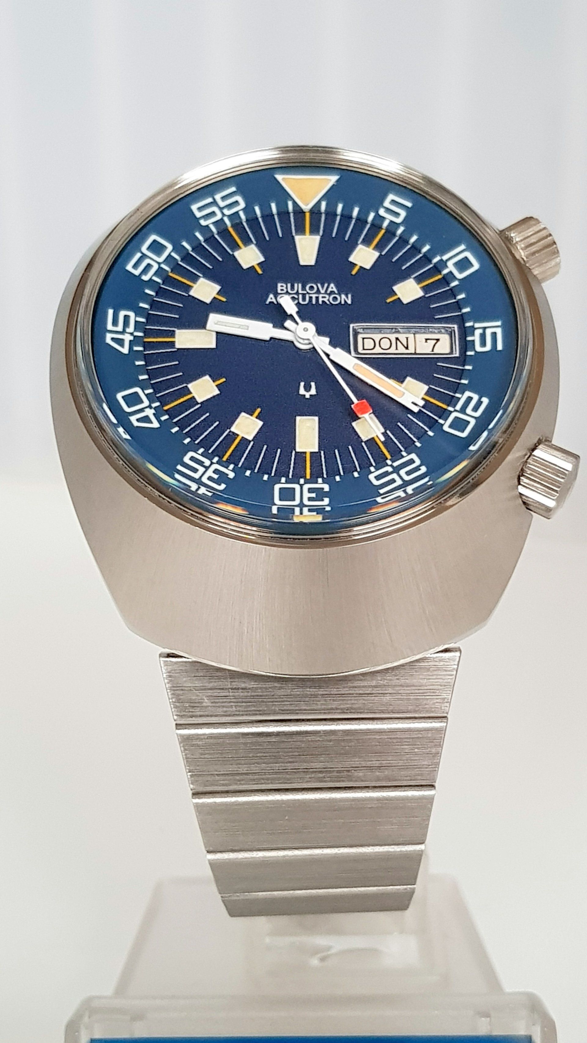Blue-dial wristwatch with silver case and band on a display stand.