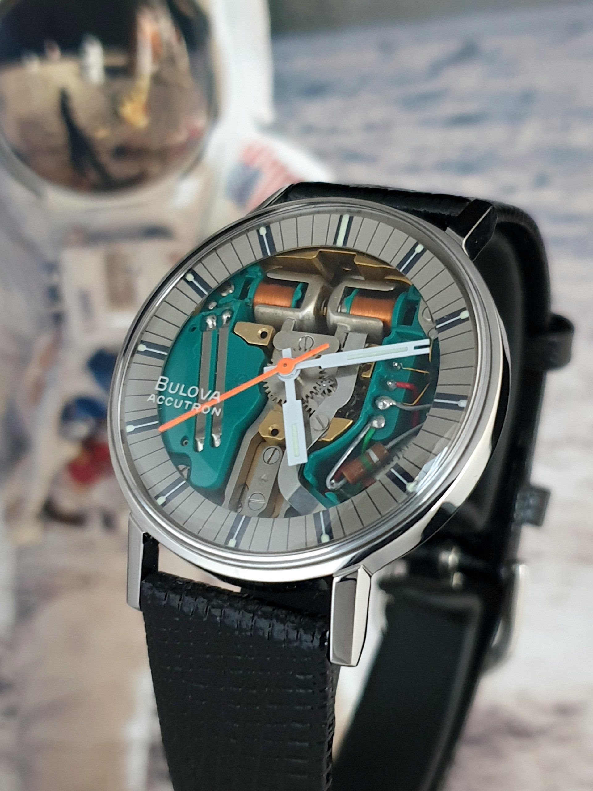 Close-up of a teal skeleton analog wristwatch with black strap on a blurred tabletop background