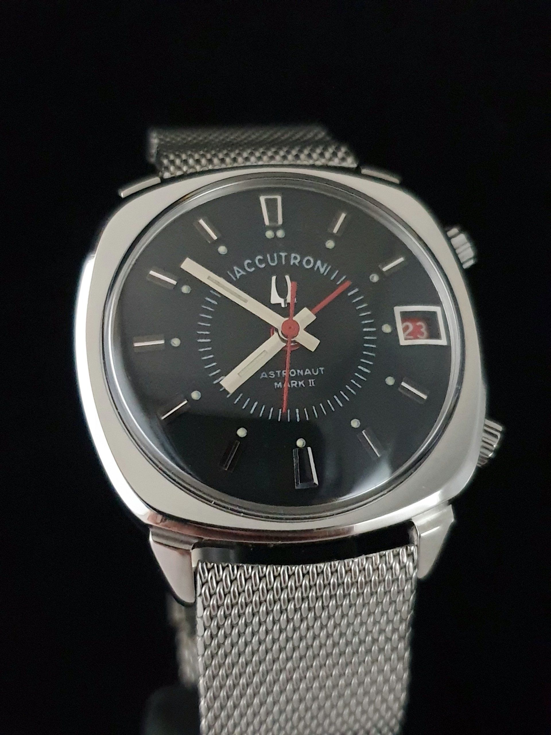 Close-up of a silver wristwatch with black dial, date window, and mesh metal band.