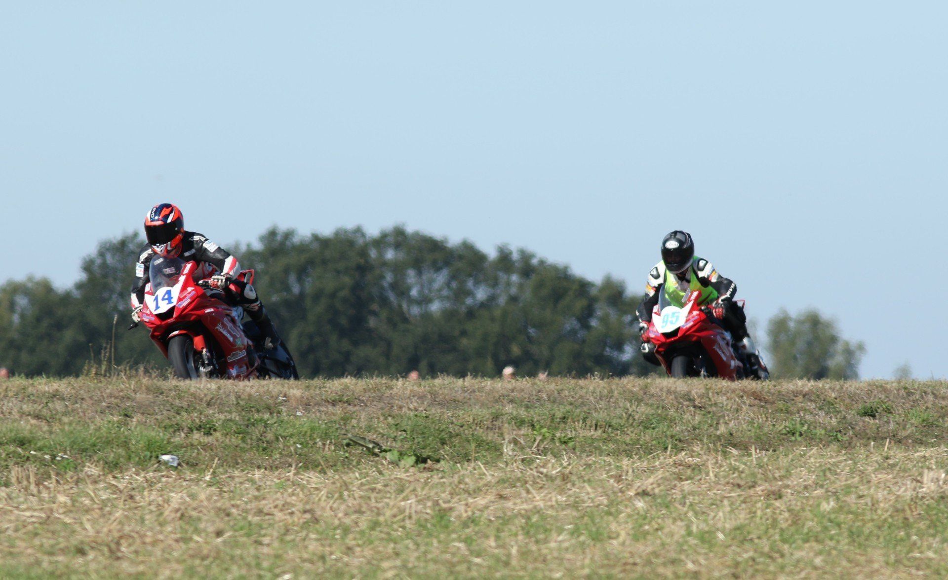 Twee rood-witte motoren racen op een circuit. De coureurs leunen op een zonnige dag in een bocht.