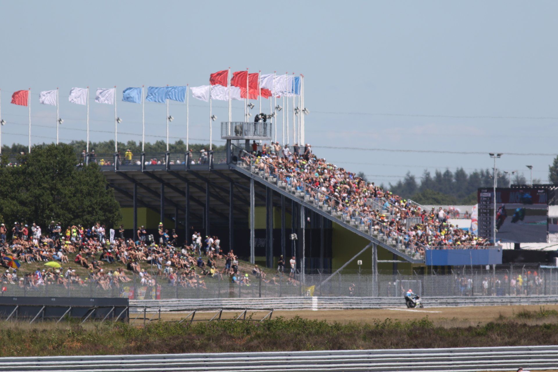 Tribunes vol toeschouwers op een racebaan. Vlaggen wapperen in de lucht en op de voorgrond rijdt een raceauto over de baan.