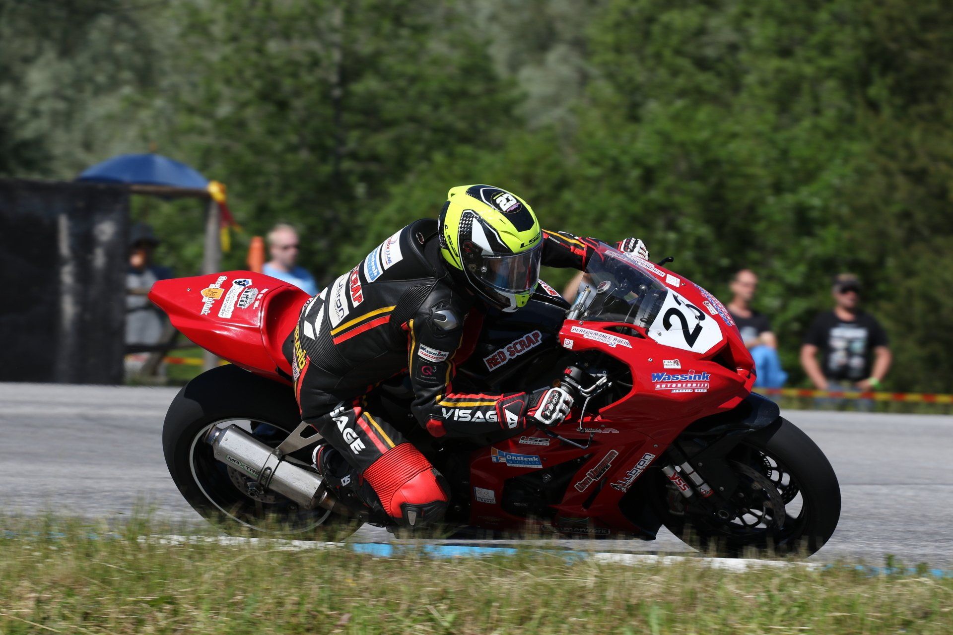Motorcoureur leunt in de bocht op een rode motor, gekleed in een zwart-rood pak en een gele helm. Het is een buitenrace.
