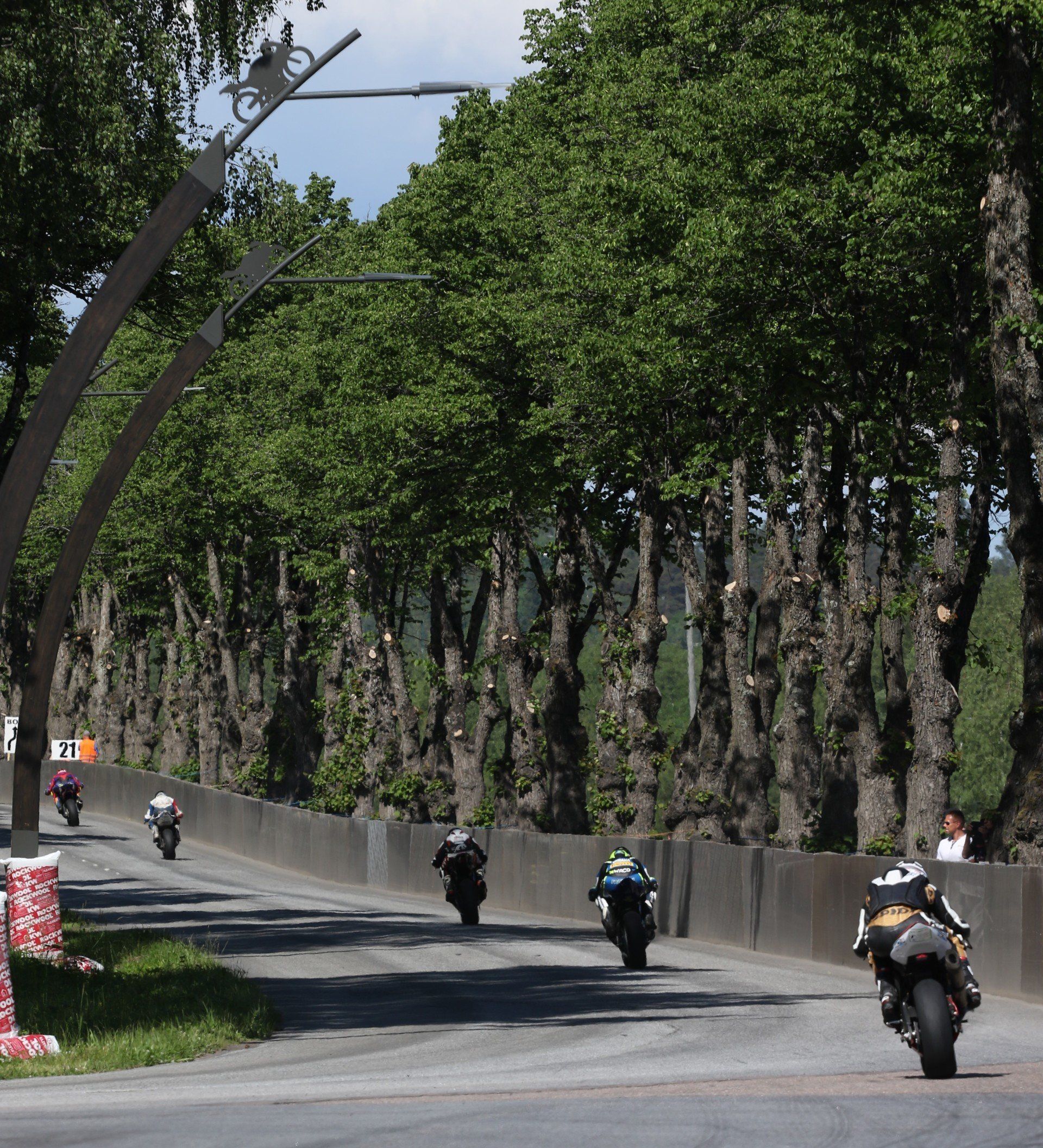 Motoren racen op een weg met bomen. Bestuurders leunen in de bochten, te zien op een zonnige dag.