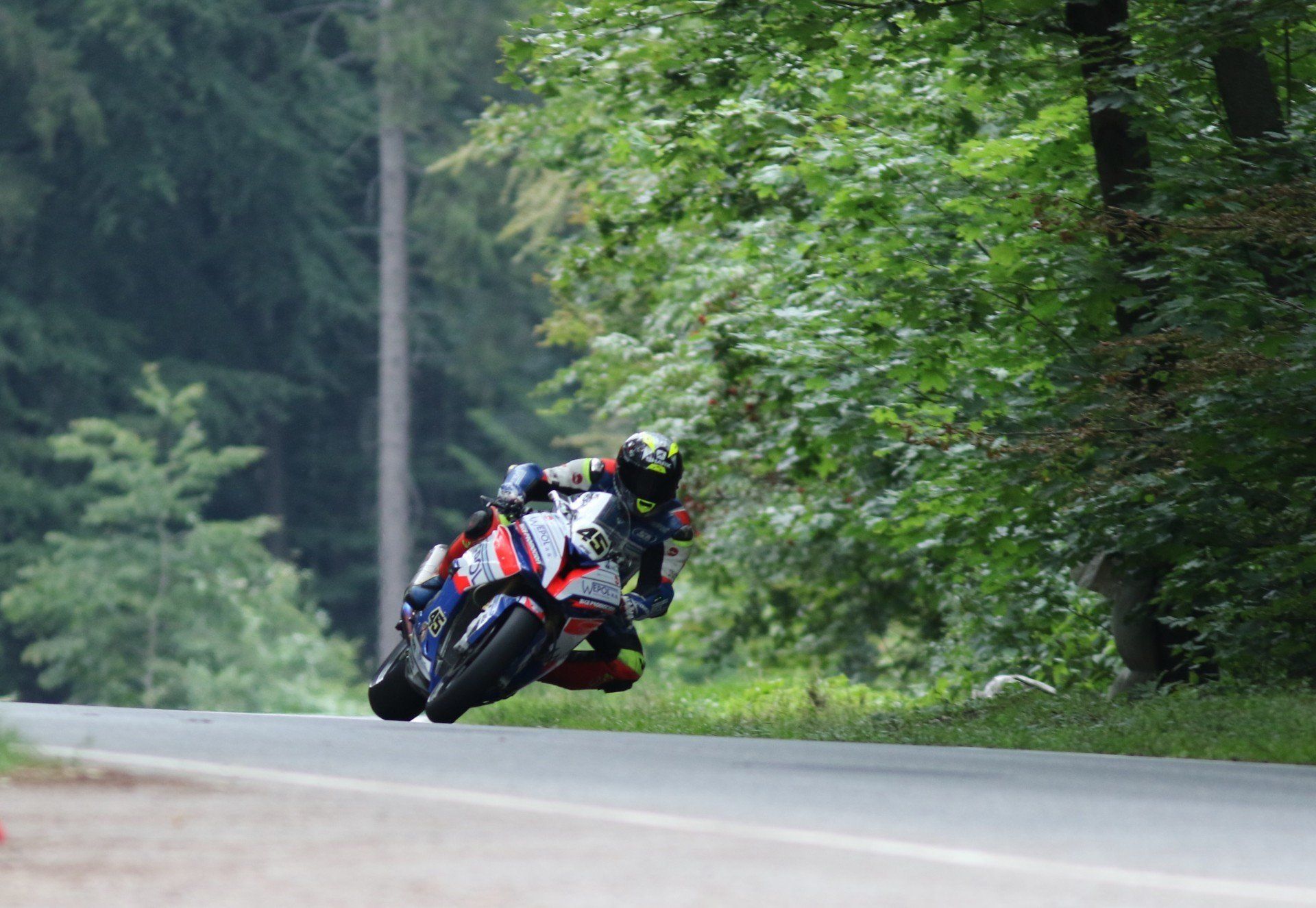 Motorrijder leunt in een bocht op een weg met bomen. De motor is rood, wit en blauw.
