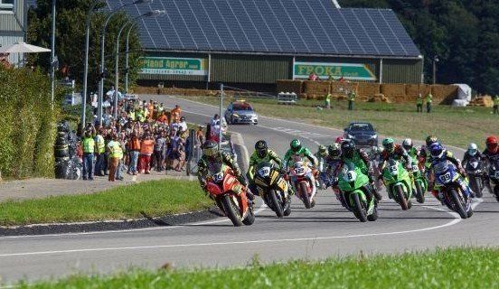 Motorrace op een verharde weg vol toeschouwers en een gebouw met zonnepanelen. De motoren zijn kleurrijk, met rijders met helmen.