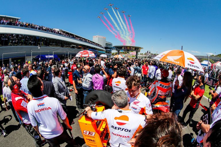 Een menigte op een racecircuit met vertegenwoordigers van Honda. Boven ons spatten kleurrijke rookwolken van straaljagers. Een stralende, zonnige dag.