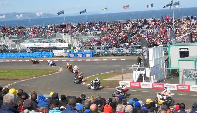 Een motorrace op een circuit, te zien vanaf de tribune vol toeschouwers. De oceaan op de achtergrond.