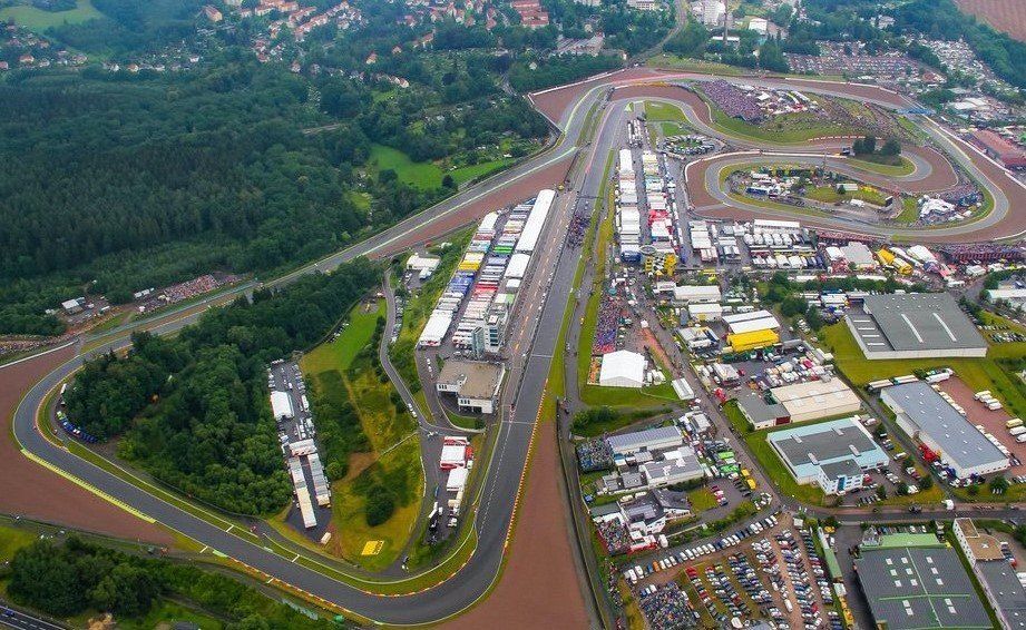 Luchtfoto van een racecircuit met pitstraat, garages en tribunes. Groene bomen omzomen het circuit; omliggende gebouwen en parkeerplaatsen zijn zichtbaar.