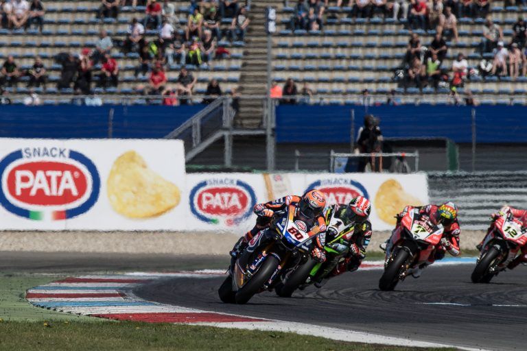 Motorcoureurs leunen in een bocht op een circuit, onder de PATA-snackreclame, toeschouwers op de achtergrond.