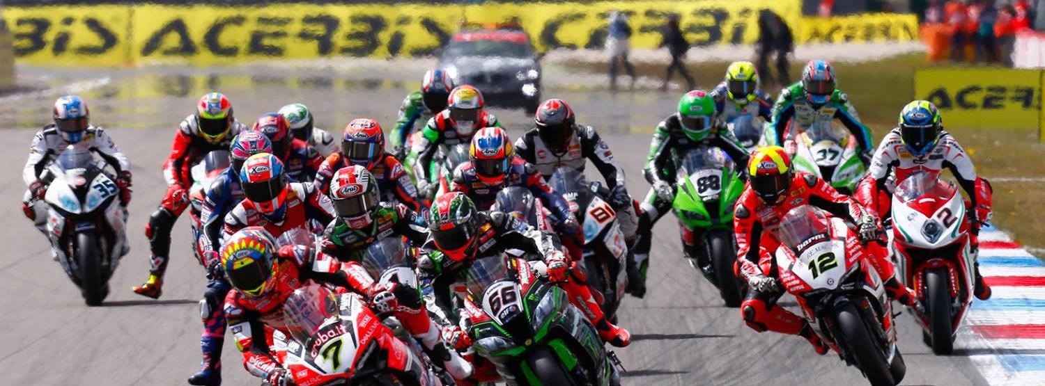 Motorrace op een circuit, veel coureurs in kleurrijke kleding leunend in een bocht. Er is een safetycar aanwezig.