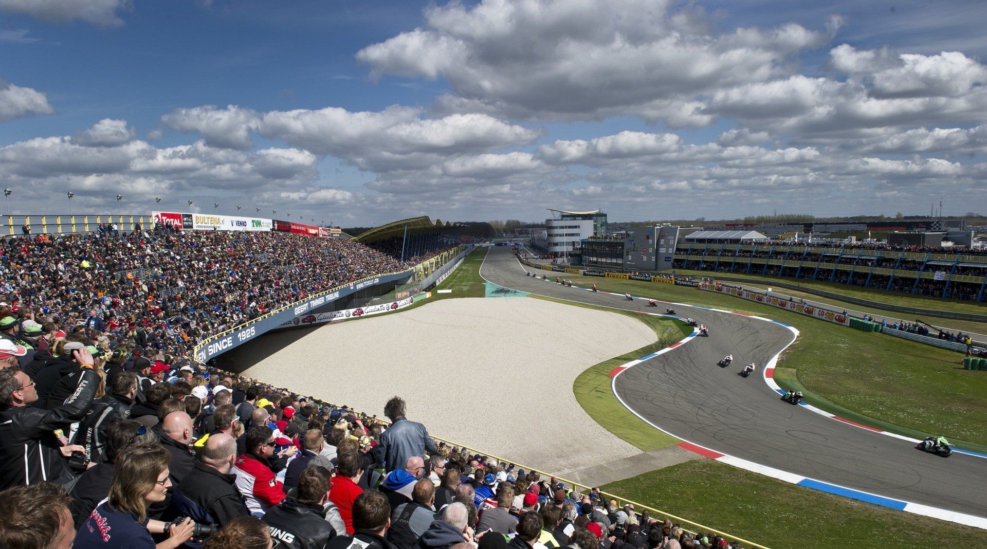 Een publiek bekijkt een motorrace op een bochtige baan op het circuit van Assen, Nederland, onder een bewolkte hemel.