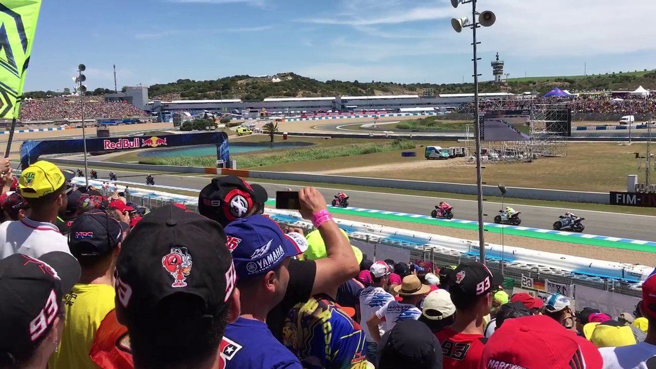 Motorrace op een circuit; toeschouwers kijken toe vanaf de tribune. Coureurs racen over het circuit; zonnige dag.
