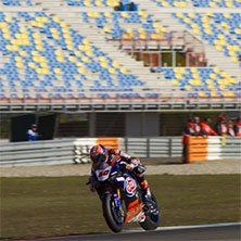 Motorcoureur op een circuit, leunend in een bocht. Blauw-geel-witte stadionstoelen op de achtergrond.