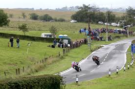 Twee motoren leunen schuin een bocht in op een natte weg tijdens een race. Toeschouwers kijken toe vanachter een hek in een landelijke omgeving.