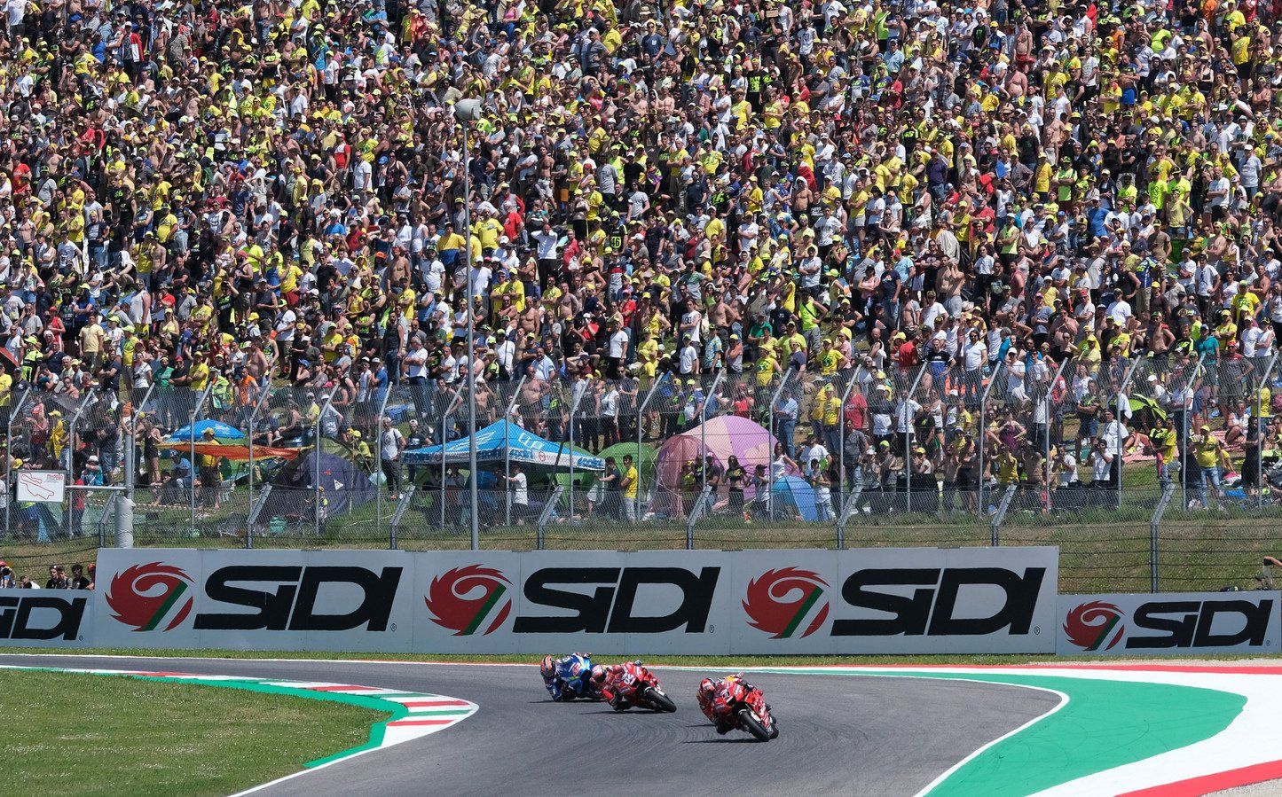 Motorrijders racen op een circuit tijdens een wedstrijd, met een groot publiek op de achtergrond.