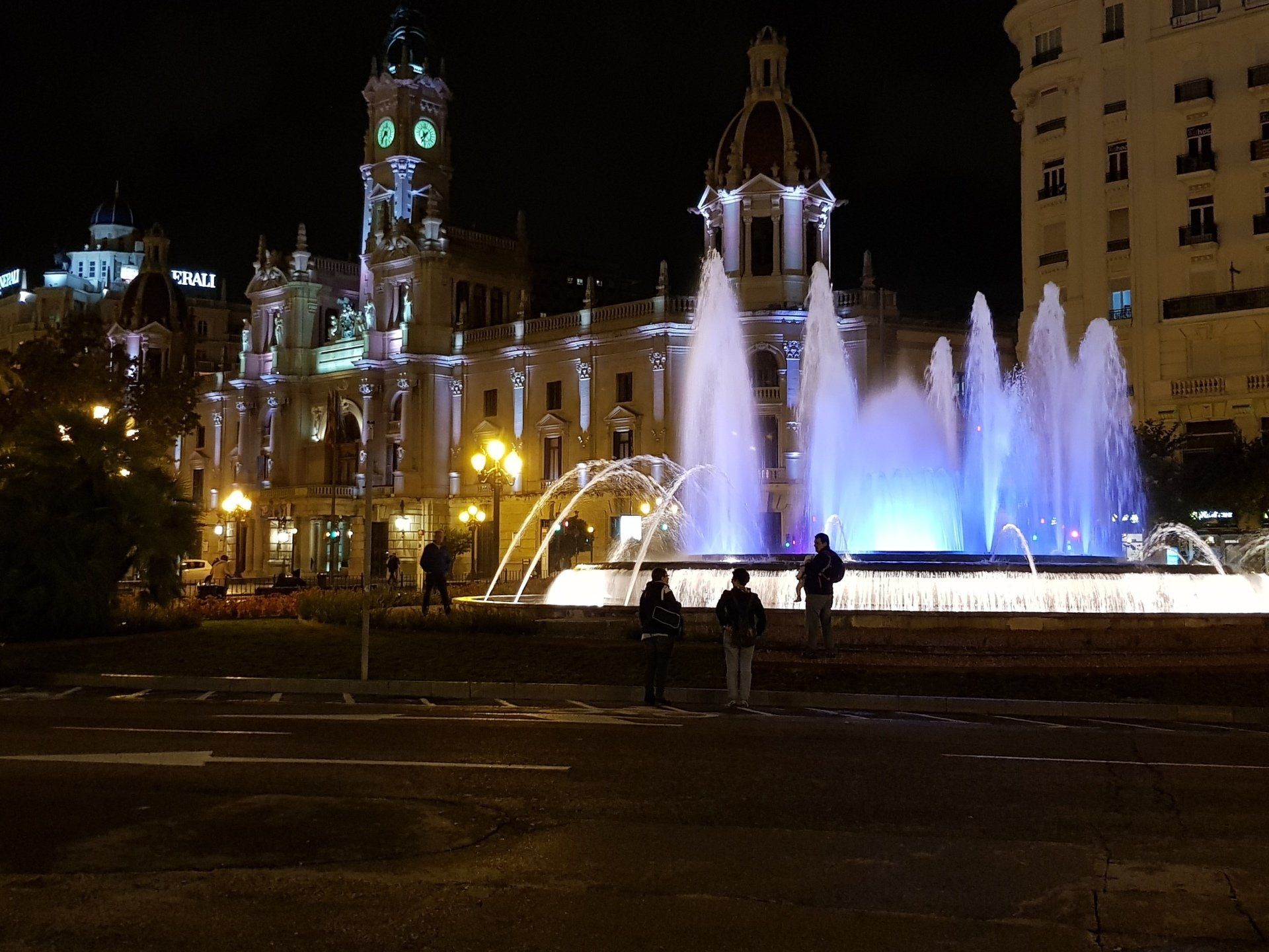 Nachtzicht op het verlichte stadhuis van Valencia en een fontein met blauw-witte lichten. Mensen lopen op het plein.