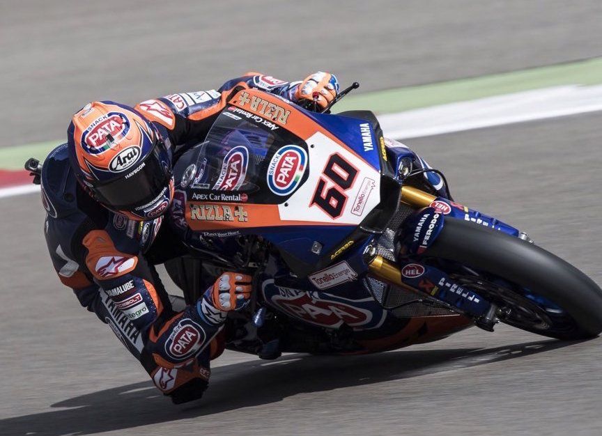Motorcoureur, gekleed in oranje en blauwe racekleding, leunt in een bocht op een racecircuit, nummer 60 is zichtbaar.
