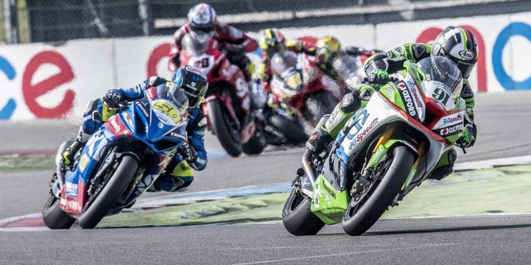 Motorcoureurs leunen een bocht in op een circuit en strijden om hun positie. Een Kawasaki met nummer 91 voert het peloton aan, met een blauwe motor vlak daarachter.