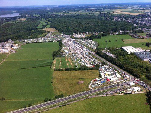 Luchtfoto van een groot buitenevenement met tenten, podia en menigten op een veld naast een snelweg en een bos.